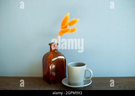 Tazza di caffè grigia, vaso di fiori secchi arancioni su tavola di legno. Sfondo blu parete Foto Stock
