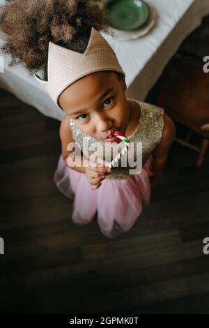 Ragazza che indossa il giocattolo corona mangiare Natale caramella canna in piedi vicino al tavolo a casa. Foto Stock