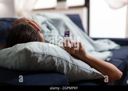 Donna malata sul divano che utilizza la tecnologia medica per il coronavirus pandemico. Adulto caucasico con pulsossimetro sul dito che misura la saturazione di ossigeno. Sintomi di Covid 19 Foto Stock