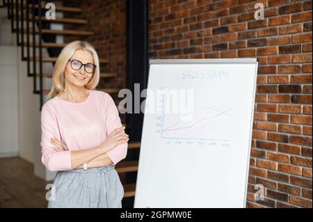 Allenatore di affari altamente qualificato con le braccia incrociate si trova vicino alla lavagna con grafici. Sorridente donna di mezza età in occhiali guarda la fotocamera, conduce un webinar online, dando formazione Foto Stock
