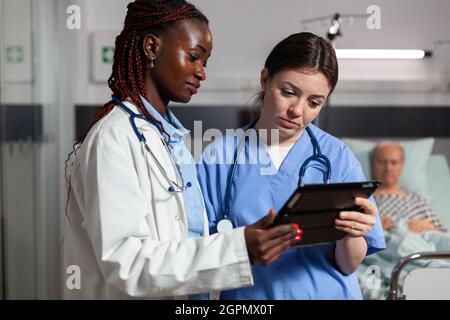 Infermiera medica che chiede consiglio dal medico afroamericano, tenendo il pc della tavoletta nella stanza dell'ospedale, circa il trattamento per l'uomo anziano malato, malato, malato che posa nel letto. Foto Stock