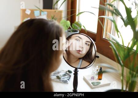 Ragazza di bellezza che guarda lo specchio mentre tocca il suo viso e l'applicazione di crema, rughe durante la routine di bellezza del mattino. Felice sorridente bella giovane donna applicando lozione idratante . Foto Stock