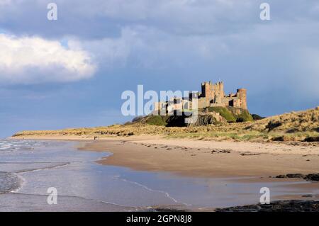 Il Castello di Bambburgh sulla spiaggia si illumina di arancione poco prima del tramonto con il cielo scuro a Bambburgh, Northumberland, Inghilterra Foto Stock