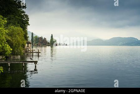 Vista sul tranquillo lago di Zugersee con montagne sullo sfondo e cielo nuvoloso, dalla città svizzera di Zug. Foto Stock