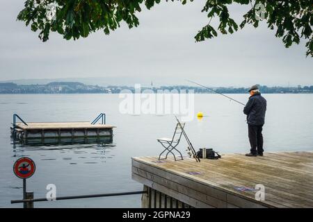 Uomo anziano che pesca tranquillamente all'alba sulle rive del Lago di Zugersee con il lago calmo e una giornata nuvolosa. Foto Stock