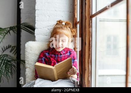 Bambina che legge un libro. Bambino sul davanzale. Grande finestra. Coperta calda. Apprendimento a distanza. Foto Stock