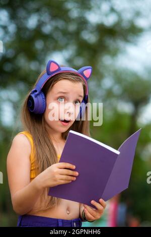 Sorpresa ragazza della scuola primaria che tiene un notebook aperto. Ragazza della scuola con libro su panca. I bambini con cuffie wireless indossano abiti alla moda, che si rilassano nel parco Foto Stock