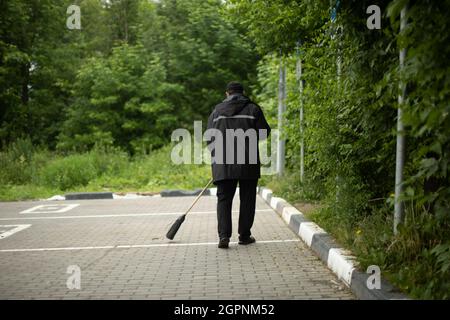 Il janitor passa la strada. Pulizia nel parcheggio. Il ragazzo pulisce lo sporco con una scopa. Mettere ordine in strada. Pulizia di Foto Stock