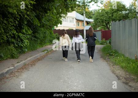 Tre ragazze stanno camminando lungo la strada. Adolescenti in campagna. Passeggiate scolari in estate. Le ragazze salgono su una collina sulla strada asfaltata. Foto Stock