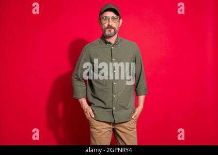 Foto di uomo età fredda indossare khaki camicia occhiali cappello isolato su sfondo rosso Foto Stock