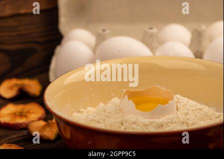 Un uovo di pollo bianco rotto in una ciotola con farina di frumento, patatine secche di mela e diverse uova intere in un vassoio di cartone bianco, primo piano selettivo Foto Stock