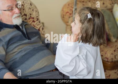 Una bambina in un cappotto medico esamina il nonno con uno stetoscopio. Giochi per bambini Foto Stock