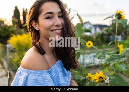 Bella giovane signora che cammina attraverso un allotment mentre spinge la sua bicicletta turchese con girasoli in un cestino. Sunbeam sullo sfondo. Foto Stock