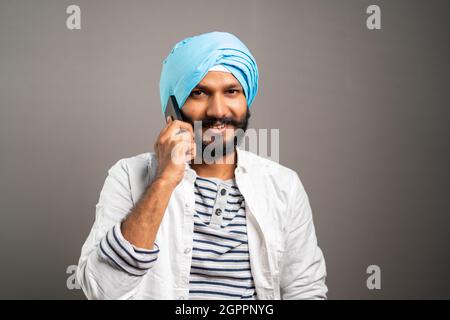 Felice sorridente indiano Sikh uomo occupato parlare sul telefono cellulare sullo sfondo studio guardando la fotocamera - concetto di comunicazione, rete e positivo Foto Stock