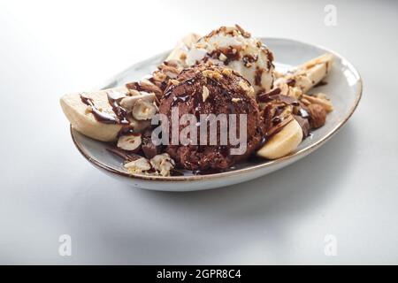 Da sopra di deliziose banane fresche servite con cioccolato e gelato alla vaniglia scoop su piatto con nocciole su tavola bianca Foto Stock