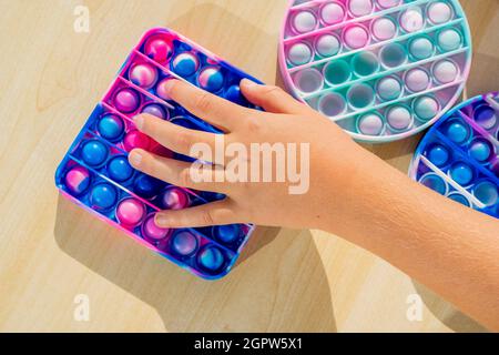 Piccolo bambino che gioca con anti stress sensoriale pop esso a tavola di legno. Mani che tengono e giocano il colore dell'arcobaleno di popit. Andamento dell'anno 2021. Vista dall'alto. Foto Stock