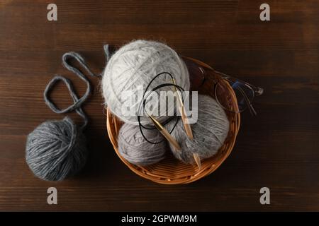 Ciotola di vimini con palle di filato e aghi da maglieria, e bicchieri su sfondo di legno Foto Stock