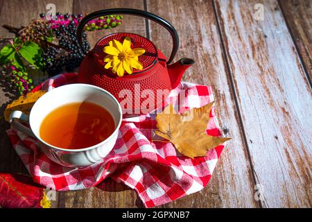 su un tavolo di legno una teiera rossa, una tazza bianca con tè, foglie d'autunno gialle e rosse, bacche nere Foto Stock