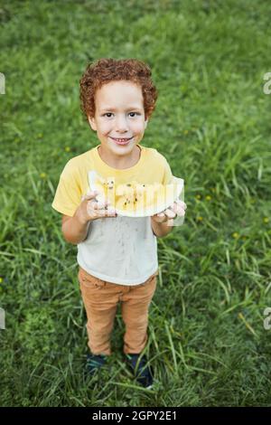 Ritratto verticale a tutta lunghezza del ragazzo riccio che tiene il cocomero all'aperto e sorride alla macchina fotografica Foto Stock