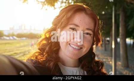 Donna con capelli rossi ricci fa sverrire selfie nel parco della città Foto Stock