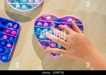 Piccolo bambino che gioca con anti stress sensoriale pop esso a tavola di legno. Mani che tengono e giocano il colore dell'arcobaleno di popit. Andamento dell'anno 2021. Vista dall'alto. Foto Stock