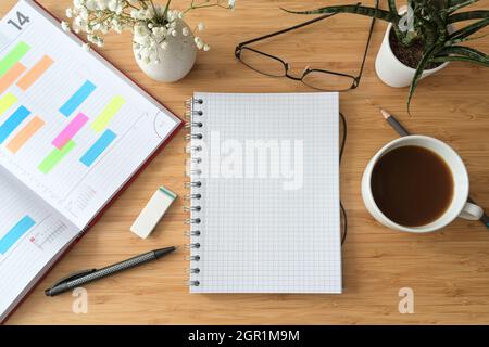 Notebook bianco a spirale su una scrivania in legno con organizer, caffè, bicchieri e fiori, spazio copia, disposizione piatta vista dall'alto, fuoco selezionato Foto Stock