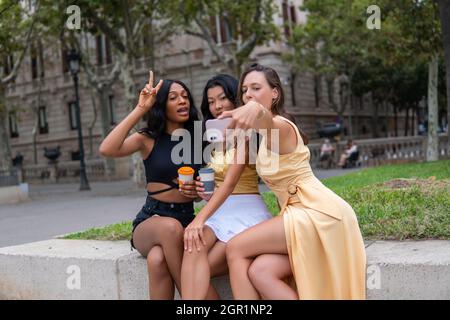 Felice giovani amici femminili diverse in abiti eleganti con bevande da asporto che prendono selfie sullo smartphone mentre si riposano insieme sulla recinzione contro vecchio edificio nella giornata estiva in città Foto Stock
