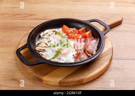 Colazione inglese in padella con uova fritte, pancetta, funghi e pomodori su stand di legno Foto Stock