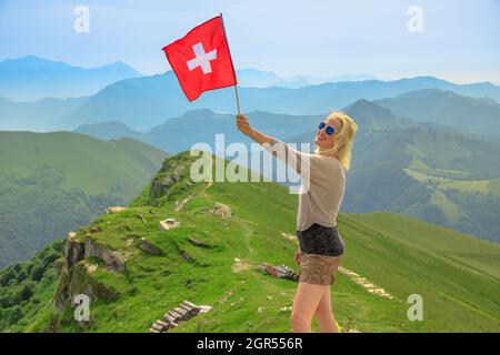 Donna turistica con bandiera svizzera sul Monte generoso Calvagione top. Vista skyline del lago di Lugano del Cantone Ticino. Stazione dei treni Top of Cog a Mendrisio Foto Stock