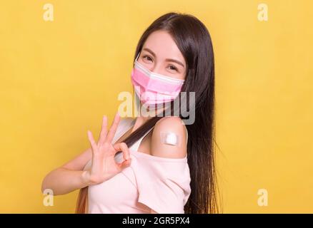 Felice giovane donna che indossa la maschera medica e mostra il suo braccio con il gesto ok dopo aver ottenuto il vaccino. Foto Stock