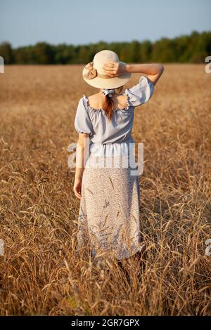 Giovane bella ragazza con capelli ricci lunghi pone in un campo di grano in estate al tramonto. Una ragazza tiene un cappello in mano contro lo sfondo di una w Foto Stock