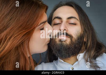 Ritratto di una giovane coppia in cui una lunga donna dai capelli rossi bacia con capelli lunghi la guancia dell'uomo dell'uomo bearded Foto Stock