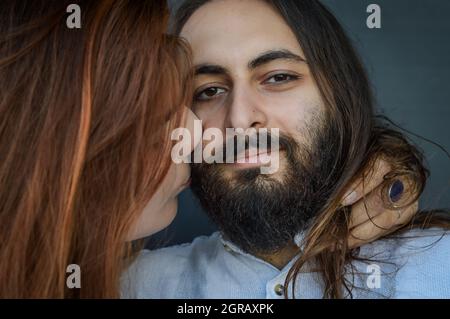 Ritratto di una giovane coppia in cui una lunga donna dai capelli rossi bacia con capelli lunghi la guancia dell'uomo dell'uomo bearded Foto Stock