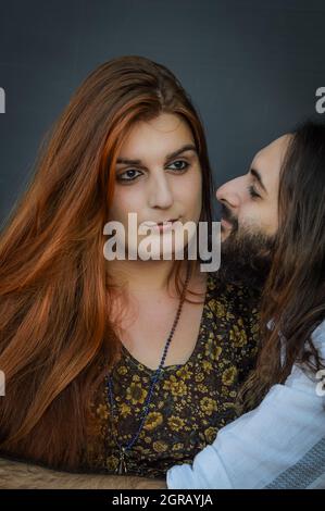 Ritratto di una giovane coppia in cui l'uomo bearded abbraccia e guarda la giovane donna dai capelli rossi mentre guarda via Foto Stock