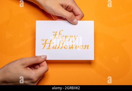 le mani femminili che tengono un pezzo di carta con l'iscrizione happy halloween in arancione scrittura su cartone di sfondo arancione Foto Stock