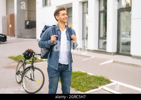 Ritratto medio di felice bel giovane uomo di consegna con grande zaino termico in piedi sulla strada della città. Foto Stock