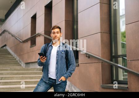 Ritratto di bell'uomo di consegna con grande zaino termico che tiene in piedi il telefono cellulare sulle scale dell'edificio dell'ufficio, guardando la fotocamera Foto Stock