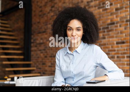 Affascinante biraciale giovane imprenditore femminile guarda la macchina fotografica, ritratto di sorridente uomo d'affari misto-gara con capelli Afro in elegante casual camicia seduta in interni, profilo aziendale Foto Stock