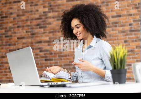 Felice donna d'affari biraciale fare videochiamata sul portatile, felice donna freelance guarda la webcam e parlare con i partecipanti online di riunione online, prendendo parte a una conferenza virtuale Foto Stock