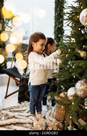 Adorabili bambine che decorano l'albero di Natale a casa, sorridendo, trascorrendo del tempo insieme. Belle sorelle che festeggiano le vacanze invernali, avendo Foto Stock