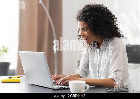 Stupito imprenditore afroamericano in camicia casual seduta alla scrivania e fissando lo schermo del laptop, sorpreso dipendente misto-gara femminile ufficio lettura inaspettata buone notizie Foto Stock