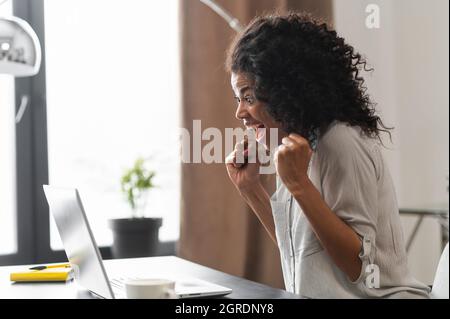 Eccitata donna d'affari afroamericana in maglietta casual seduta alla scrivania di fronte al laptop e celebrare la vittoria, sorprende dipendente di ufficio misto gara che solleva pugni in un gesto trionfo Foto Stock