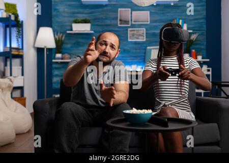 Coppia interrazziale che gioca con occhiali e controller vr per il divertimento a casa. Donna afroamericana che indossa occhiali, cuffie mentre si gode il video gioco sulla console TV con tecnologia. Foto Stock
