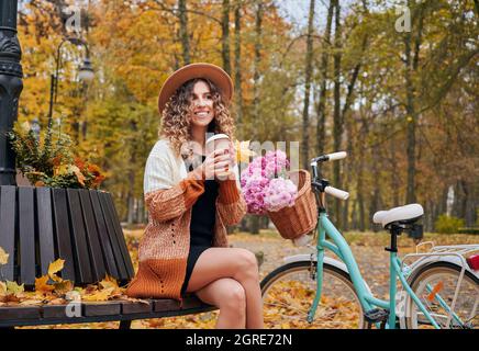 Pausa caffè mattutina nel parco cittadino con bicicletta in autunno. Simpatica ragazza seduta su panca rotonda all'aperto con la bicicletta della città delle donne ammirando il paesaggio autunnale. Foto Stock