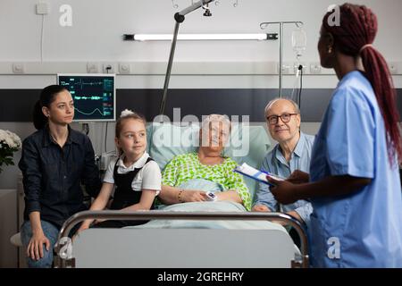 Infermiera afroamericana che parla di sintomi di malattia con anziana anziana anziana malata in reparto ospedaliero. Famiglia allegra in visita paziente in pensione che la sostiene durante il recupero clinico. Terapia medica Foto Stock