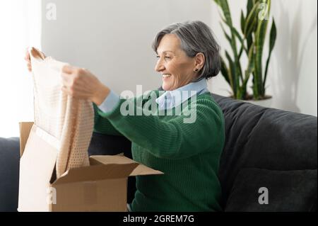 Donna adulta anziana felice sta guardando il suo nuovo jumper beige, seduto sul divano, e disimballaggio pacco. Sorridente signora online che apre online negozio ordine nella casella di spedizione postale consegna Foto Stock