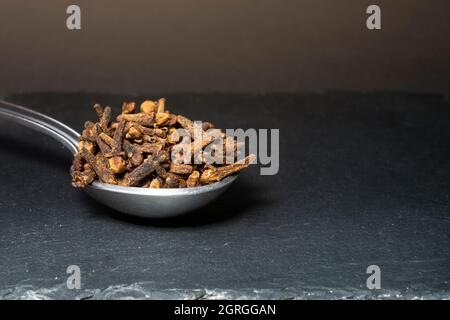 cucchiaino di metallo pieno di chiodi di garofano, isolato su sfondo scuro, tessuto di pietra nera, macro shot Foto Stock