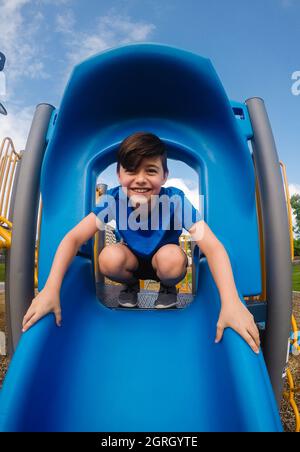 Felice scuola ragazzo vecchio seduto in cima a uno scivolo nel parco giochi. Foto Stock