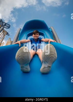 Vista ad angolo basso del ragazzo seduto in cima a uno scivolo nel parco giochi. Foto Stock