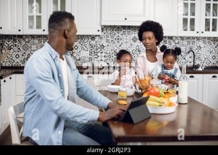 Donna afroamericana che fa colazione con due simpatiche figlie mentre marito lavora su un tablet digitale. Mattina di famiglia felice in cucina luminosa. Foto Stock